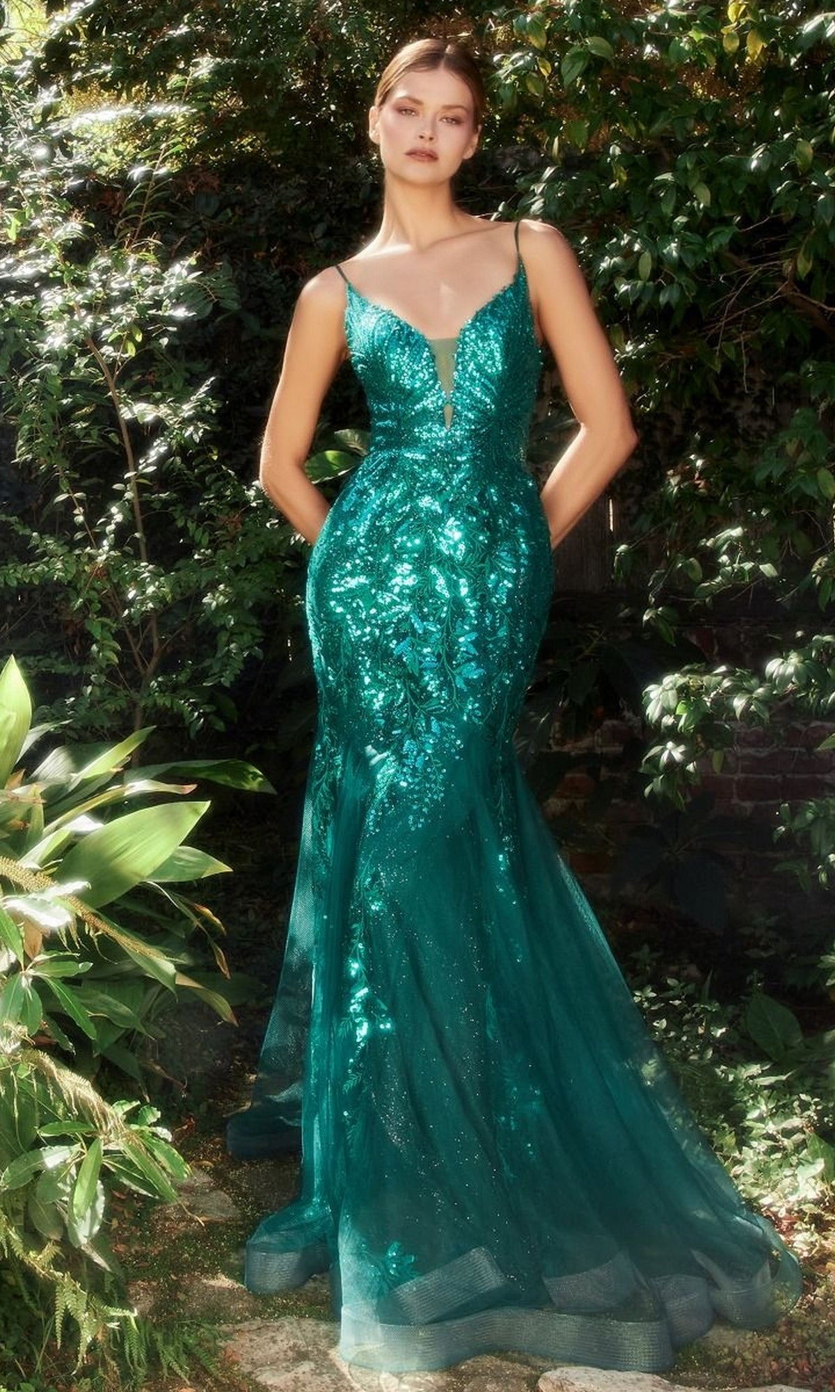 A woman is outdoors wearing the Sequin-Print V-Neck Long Mermaid Prom Dress A1118, a fitted emerald green gown featuring thin straps, sequins, and a deep V neckline.